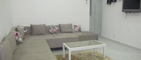 A living room with a beige sofa, a coffee table, and a patterned rug. There are three framed pictures on the wall and a television mounted on the wall.