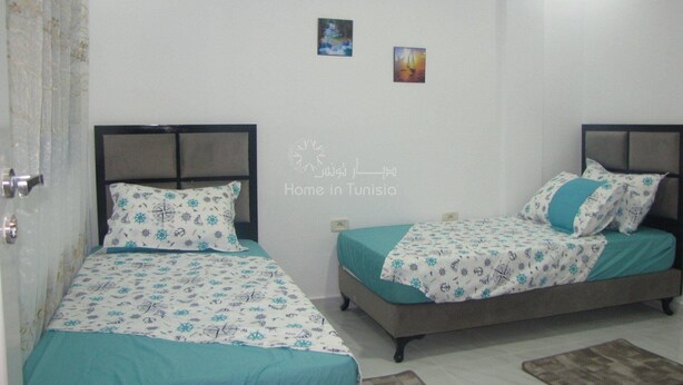 Un dormitorio con dos camas, cada una con ropa de cama y almohadas estampadas, una pared blanca con un letrero que dice 'Home in Tunisia' y una puerta con cortina.