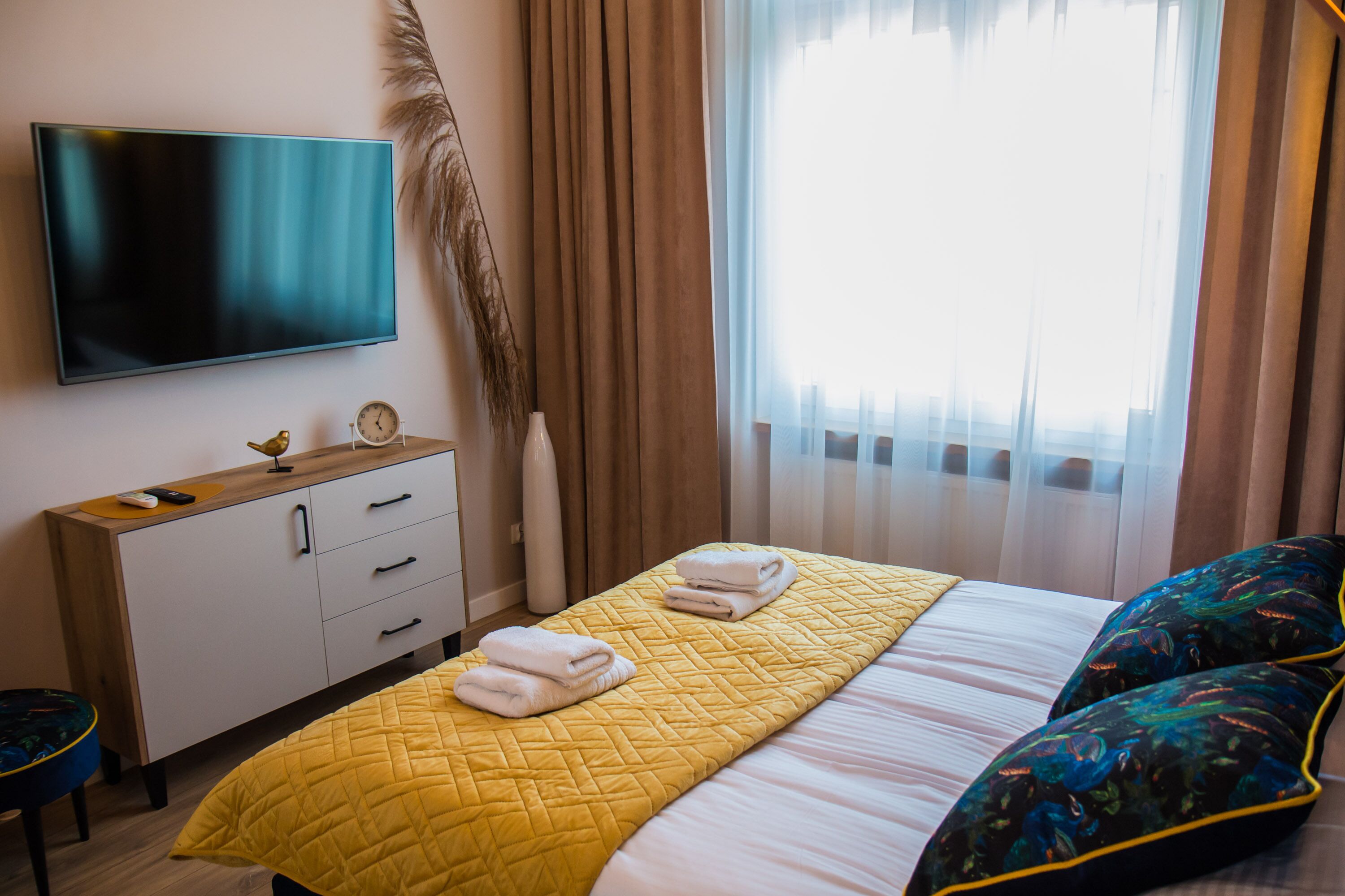 A hotel room with a bed, a TV mounted on the wall, a bedside table with a lamp, and a window with curtains.