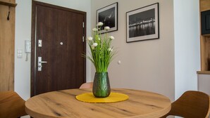 A round wooden dining table with a yellow placemat, surrounded by wooden chairs. A green vase with white flowers is placed on the table. - Miedziany (Torun)