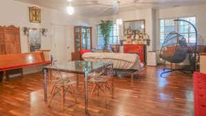 A spacious living area with a wooden floor, a glass dining table with chairs, a sofa, a hanging chair, and a large window providing natural light.