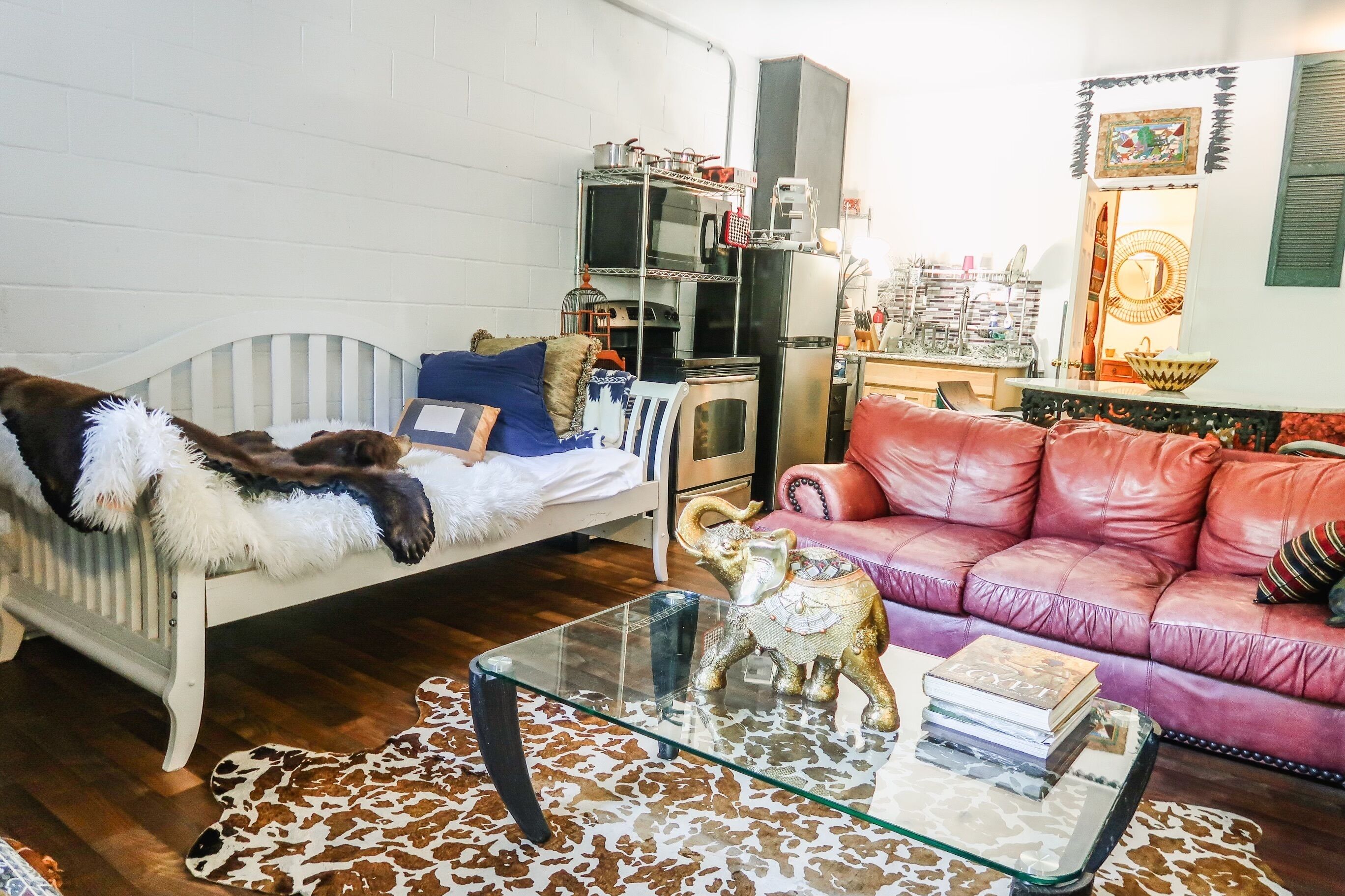 Un salon avec un canapé blanc, un divan en cuir rouge, une table basse en verre agrémentée d’objets décoratifs, un radiateur blanc et un coin cuisine équipé d ’appareils électroménagers.