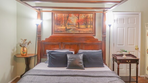 A bedroom with a four-poster bed, a painting of a forest, a bedside table with a vase, and a door with a decorative panel.