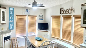 Ein Essbereich mit einem Holztisch, weißen Stühlen und einer Bank. Ein Fernseher steht auf einem Schrank und über dem Tisch hängt ein Deckenventilator.