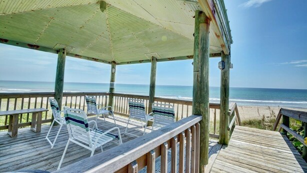 Eine Holzterrasse mit überdachtem Bereich, Stühlen und einer Bank mit Blick auf den Strand.