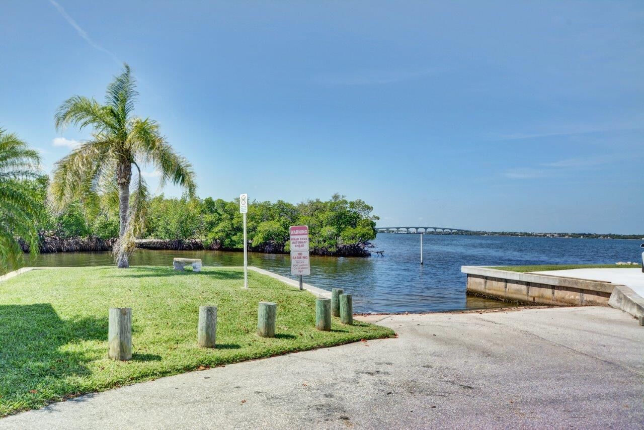 A waterfront park with a concrete walkway, palm trees, and a warning sign about no parking.