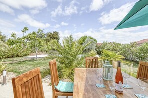 An outdoor dining area with wooden chairs and a table, surrounded by greenery and a clear sky.