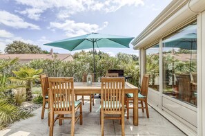 An outdoor patio with a wooden table and chairs, a green umbrella, and a glass door leading to a garden.