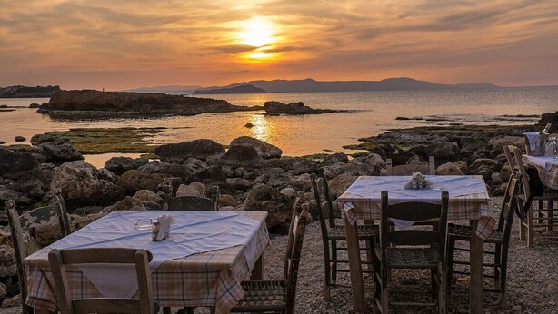 A seaside restaurant with tables and chairs set up for dining, overlooking a rocky coastline and the sea during sunset. - An ambient 19th century semi-basement studio only steps from the Cretan Sea (Χανιά)