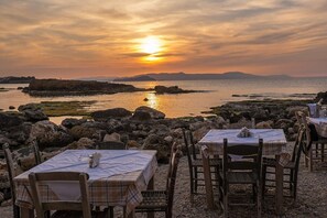 A seaside restaurant with tables and chairs set up for dining, overlooking a rocky coastline and the sea during sunset. - An ambient 19th century semi-basement studio only steps from the Cretan Sea (Χανιά)
