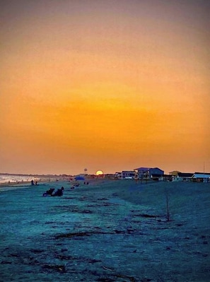 Une scène de plage avec un coucher de soleil, des maisons et une jetée en arrière-plan.