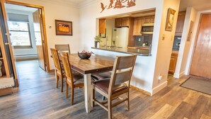 Un coin repas avec une table et des chaises en bois, une cuisine à l’arrière-plan et une porte donnant sur une autre pièce.