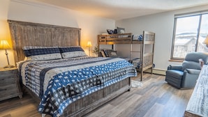 Une chambre avec un grand lit, un lit superposé, un bureau avec une chaise et une fenêtre donnant sur un beau paysage.