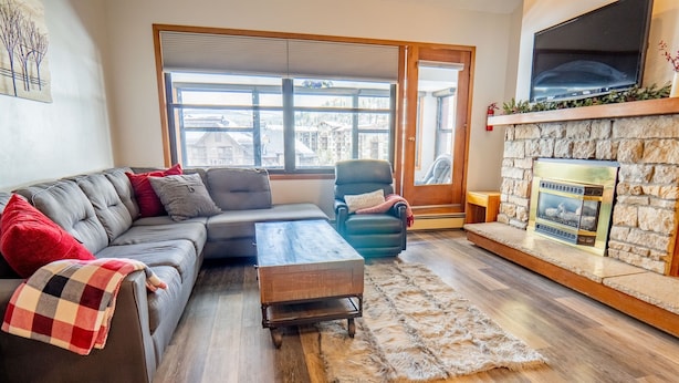 Un salon comprenant un canapé d’angle, un petit fauteuil, une table basse en bois, une cheminée en pierre et une grande fenêtre donnant sur des immeubles.
