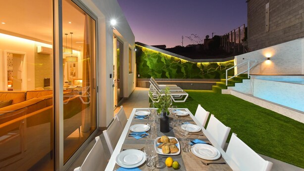 An outdoor dining area with a table set for four, surrounded by a modern house with glass doors and a green wall.