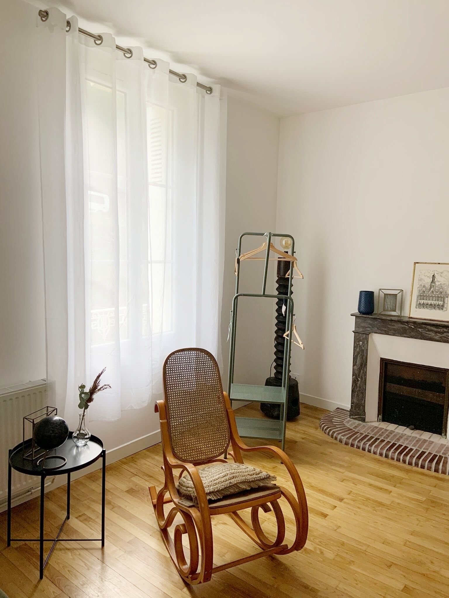 Un salon comprenant un fauteuil à bascule en bois, une petite table avec un vase, une étagère en métal et une cheminée.