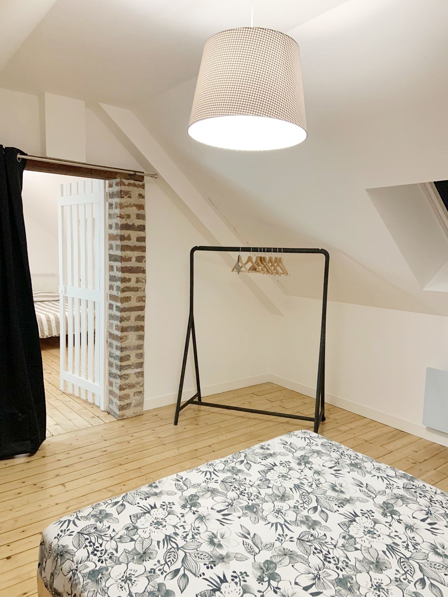 Une pièce avec un plafond en pente, un luminaire suspendu, un portant à vêtements en métal, un lit avec une literie à motifs floraux et un mur en briques.