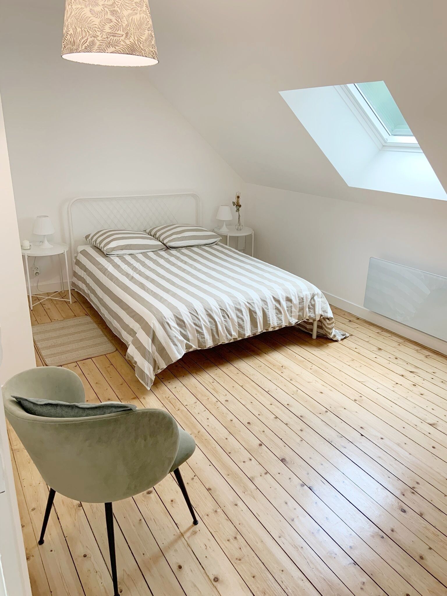 Quarto com cama, cadeira, mesa de cabeceira, luminária e claraboia.