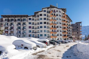 Un hotel de varios pisos con balcones, un estacionamiento con coches cubiertos de nieve y montañas al fondo.