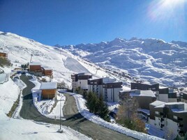 Un pueblo en la montaña cubierto de nieve con edificios modernos, un camino serpenteante y un cielo azul despejado.