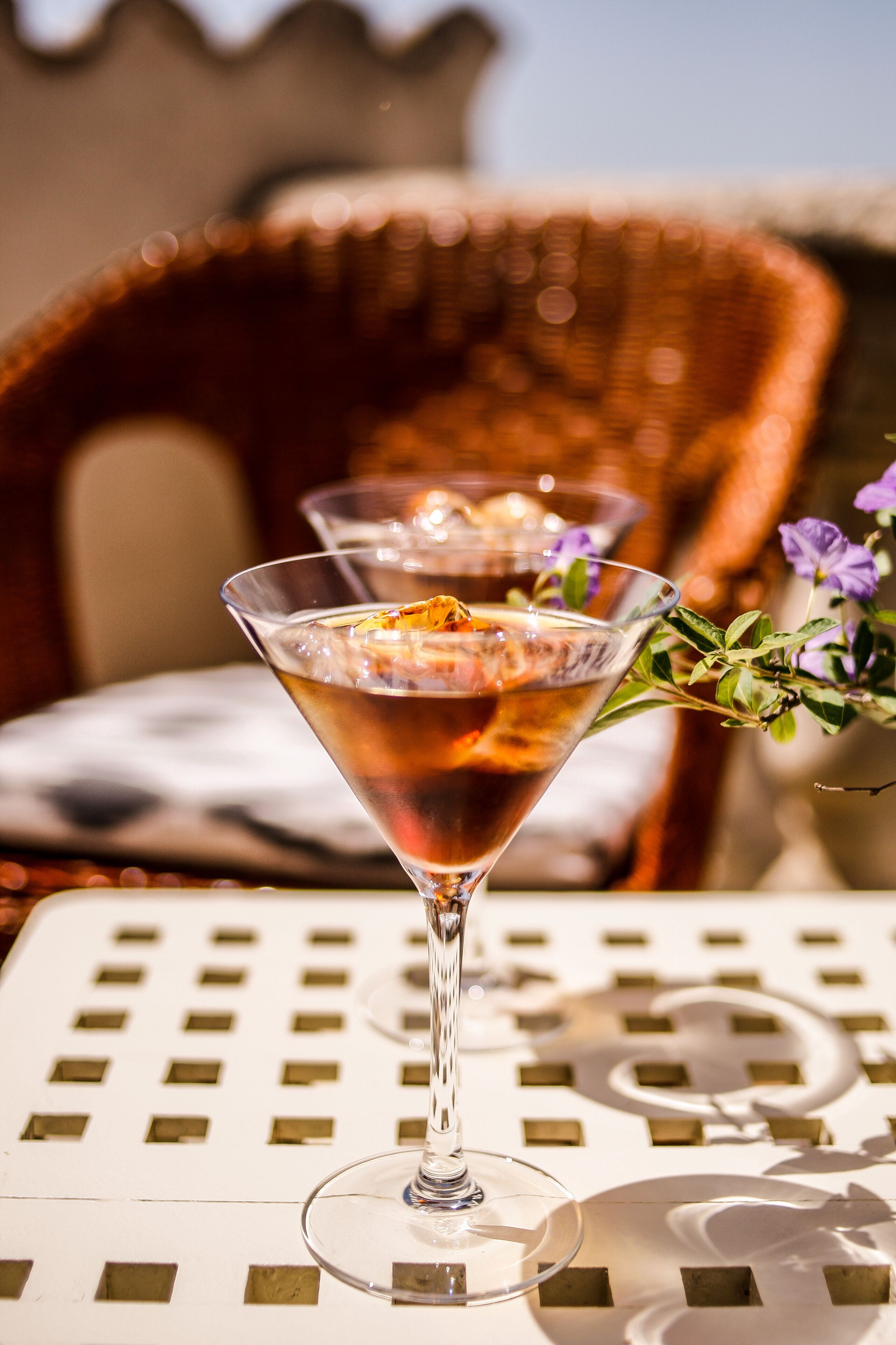 Two martini glasses on a table with a patterned tabletop, a wicker chair in the background, and a small branch with purple flowers.