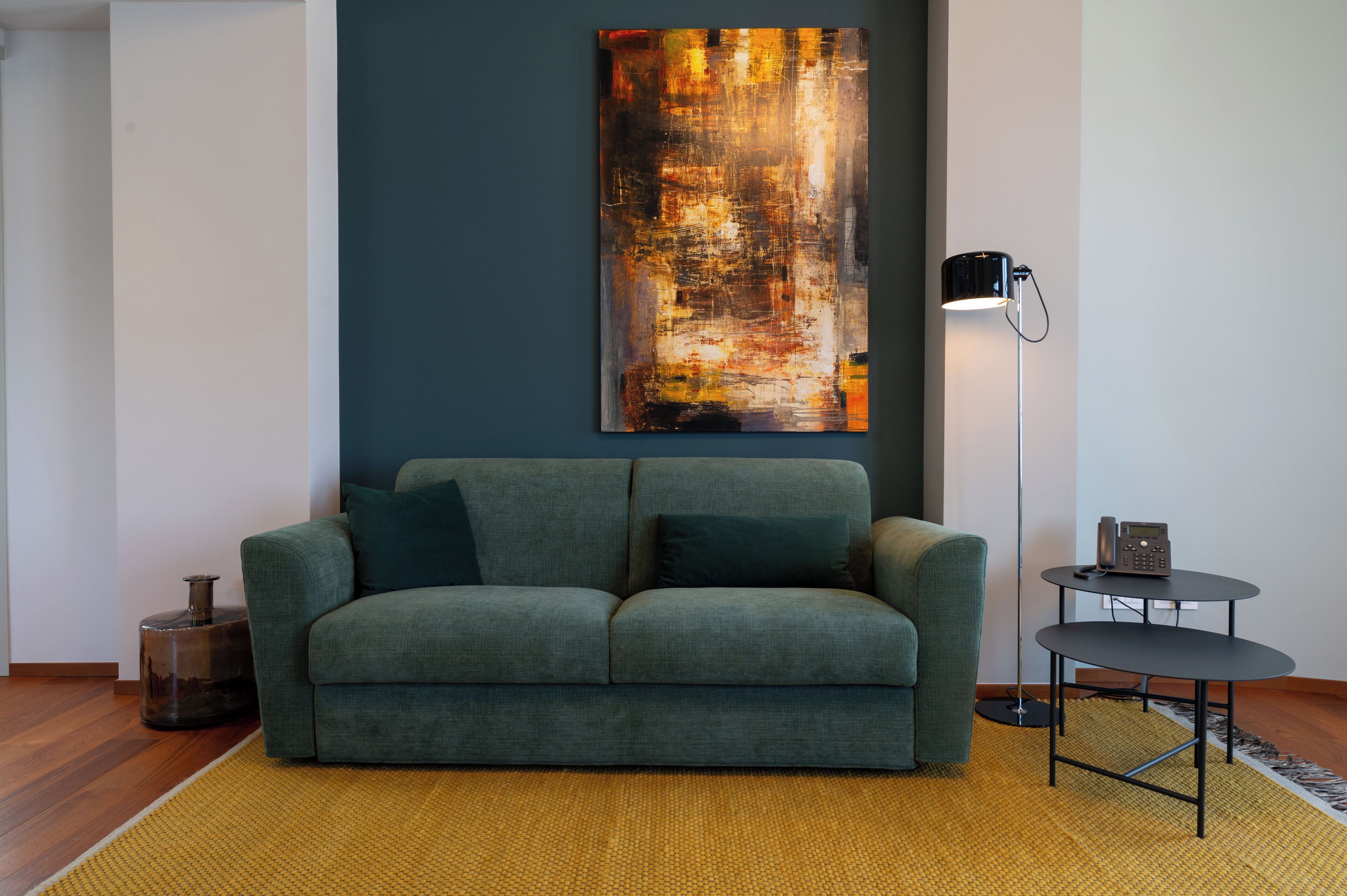 Ein modernes Wohnzimmer mit einem grünen Sofa, einem großen abstrakten Gemälde an der Wand, einer Stehlampe und einem kleinen Tisch mit einem Telefon.