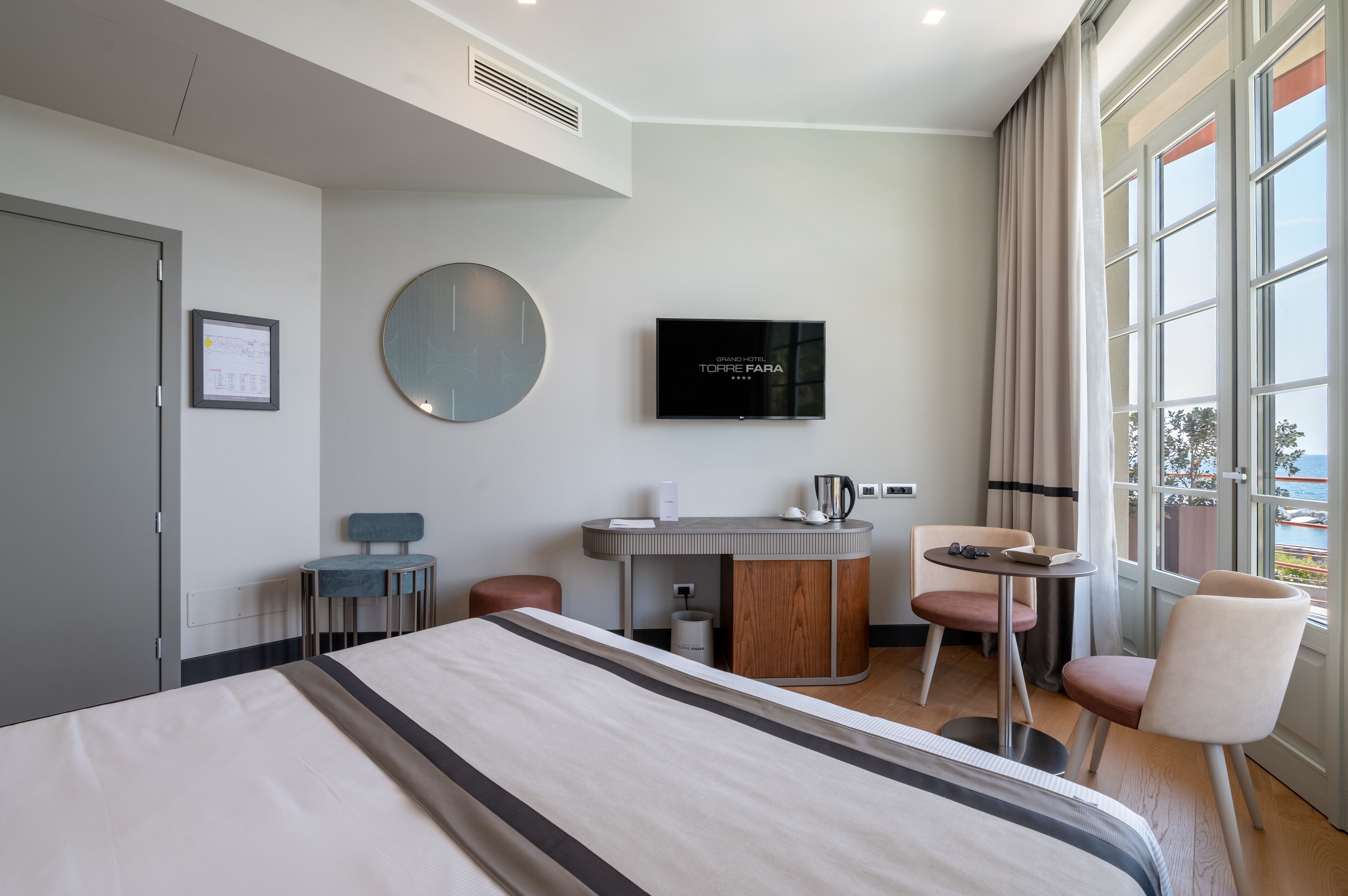 A modern hotel room with a large bed, a desk, a TV, and a view of the sea.