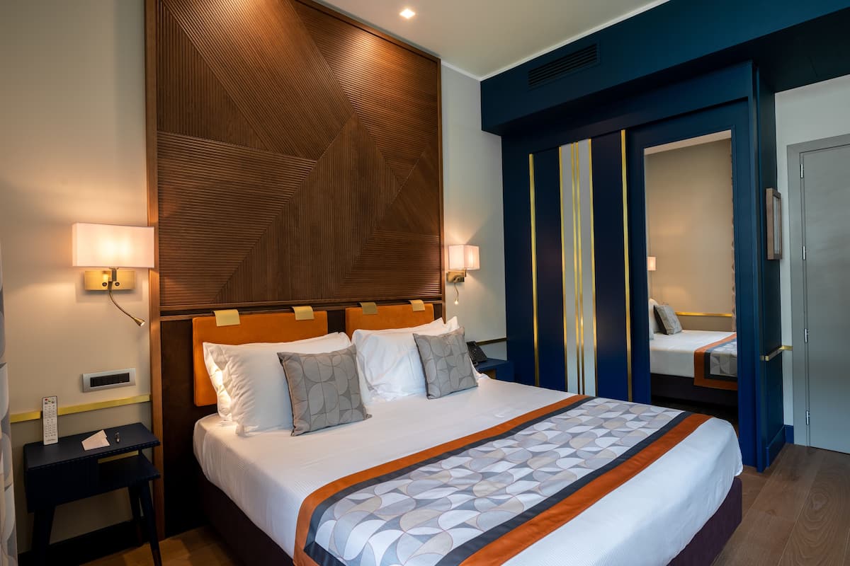 A hotel room with a large bed, bedside lamps, a wooden headboard, a wardrobe, and a small desk with a lamp.