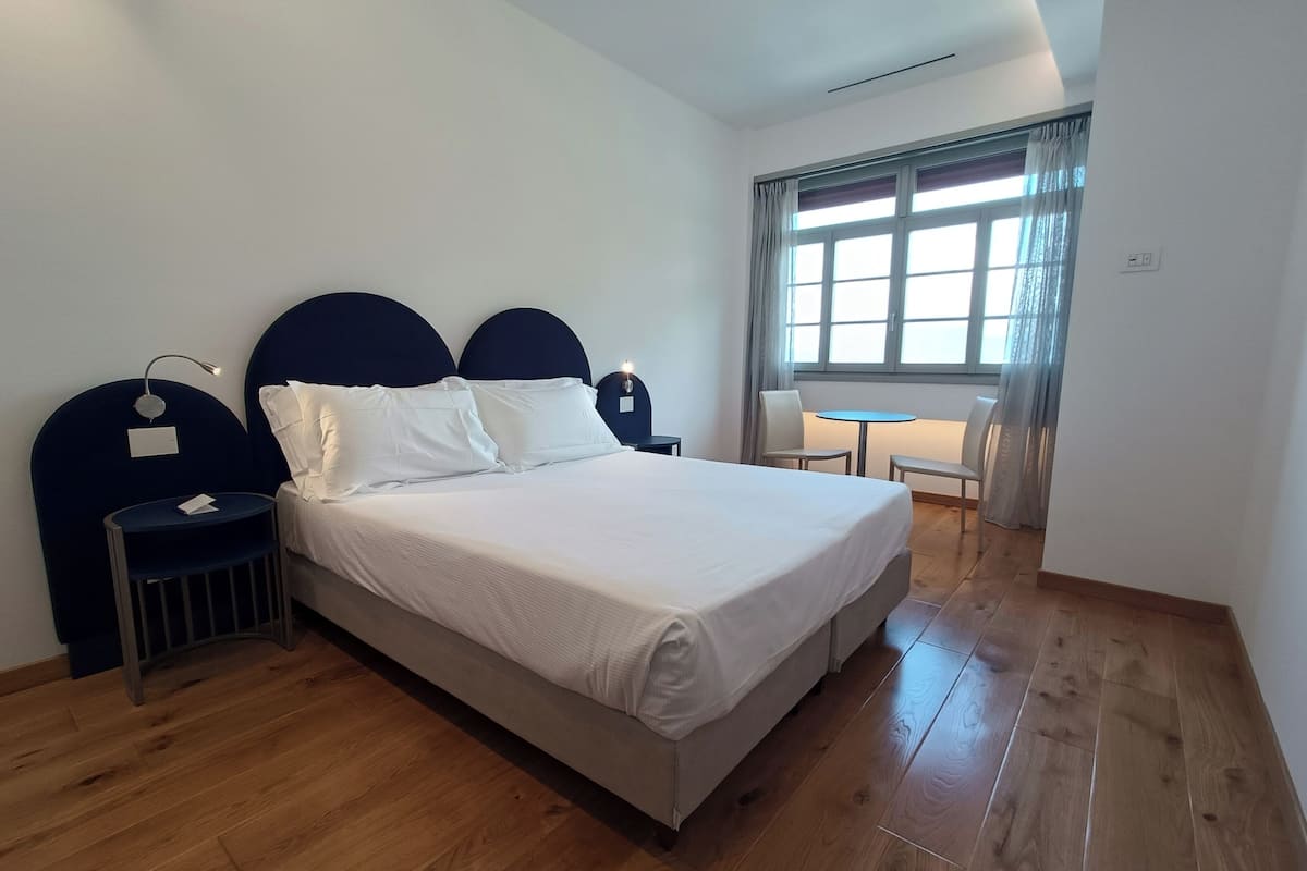A modern hotel room with a large bed, a bedside table, a nightstand, a window with curtains, and a small table with chairs.