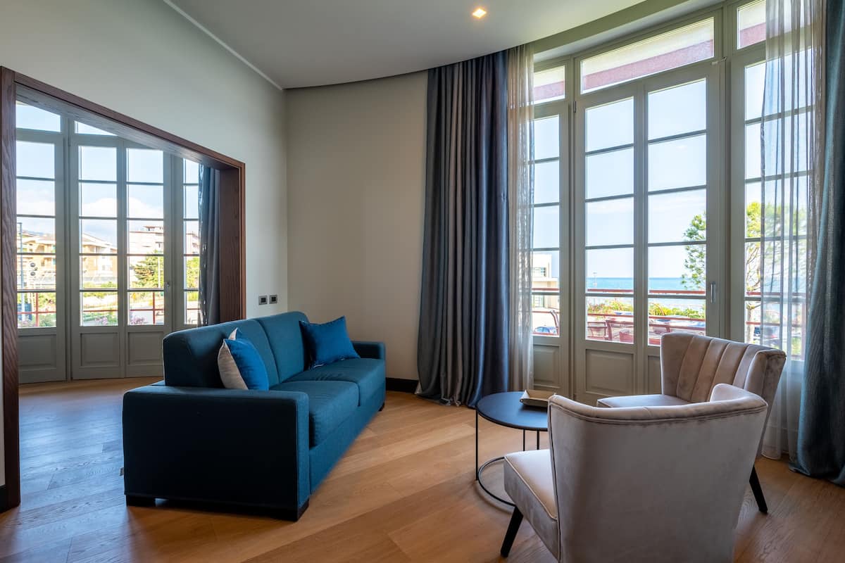 A modern living room with a blue sofa, a white armchair, and a small round table. Large windows with curtains provide a view of the outside.