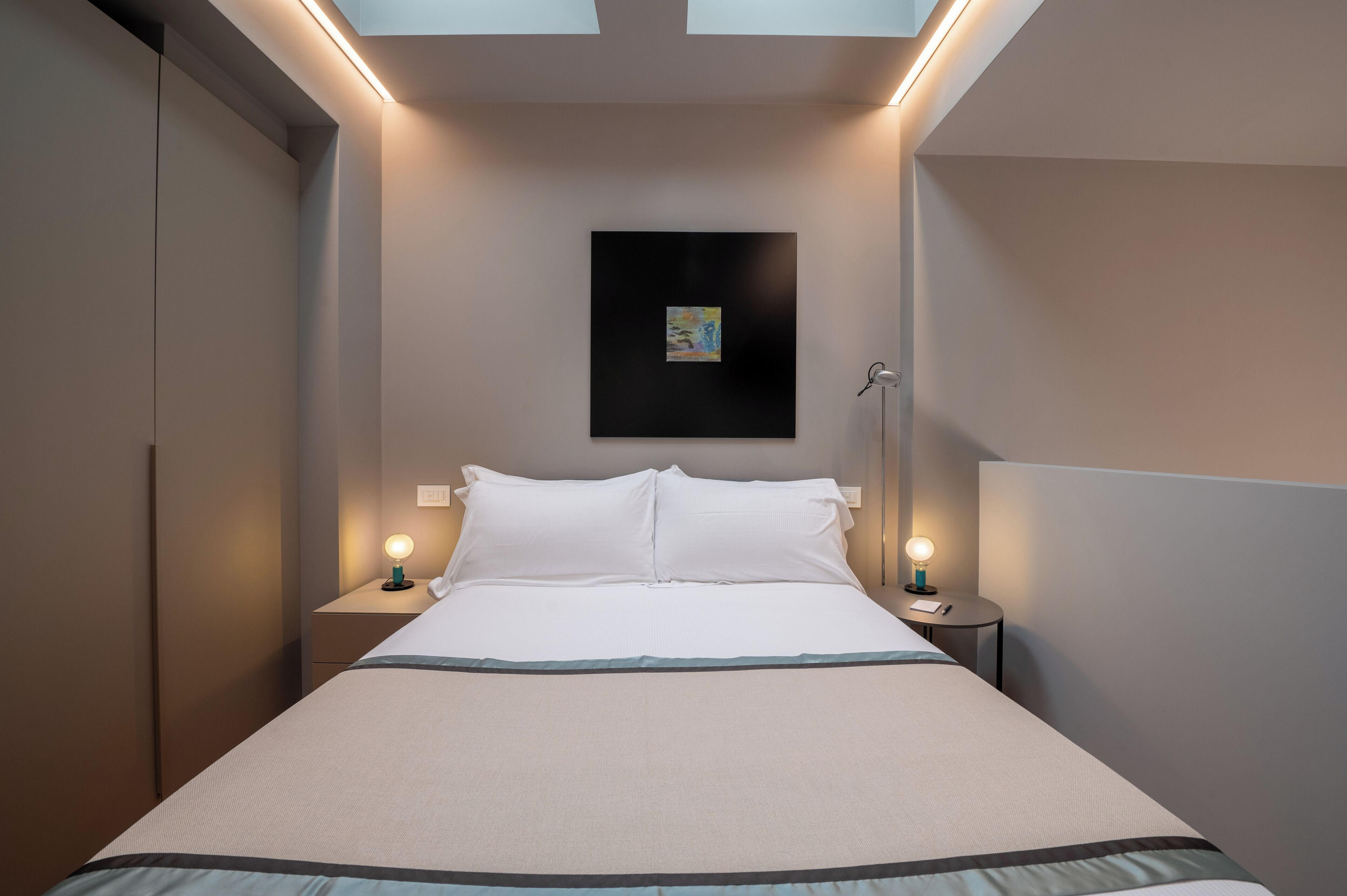 Ein ordentlich bezogenes Bett mit weißen Leinentextilien, zwei Nachttische mit Lampen, ein an der Wand hängendes Kunstwerk und eine Deckenleuchte.