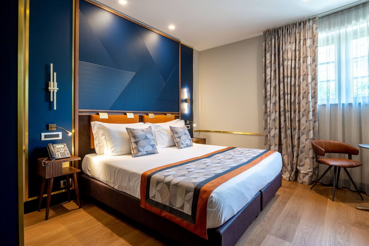 A hotel room with a large bed, a bedside table with a phone, and a chair by the window.