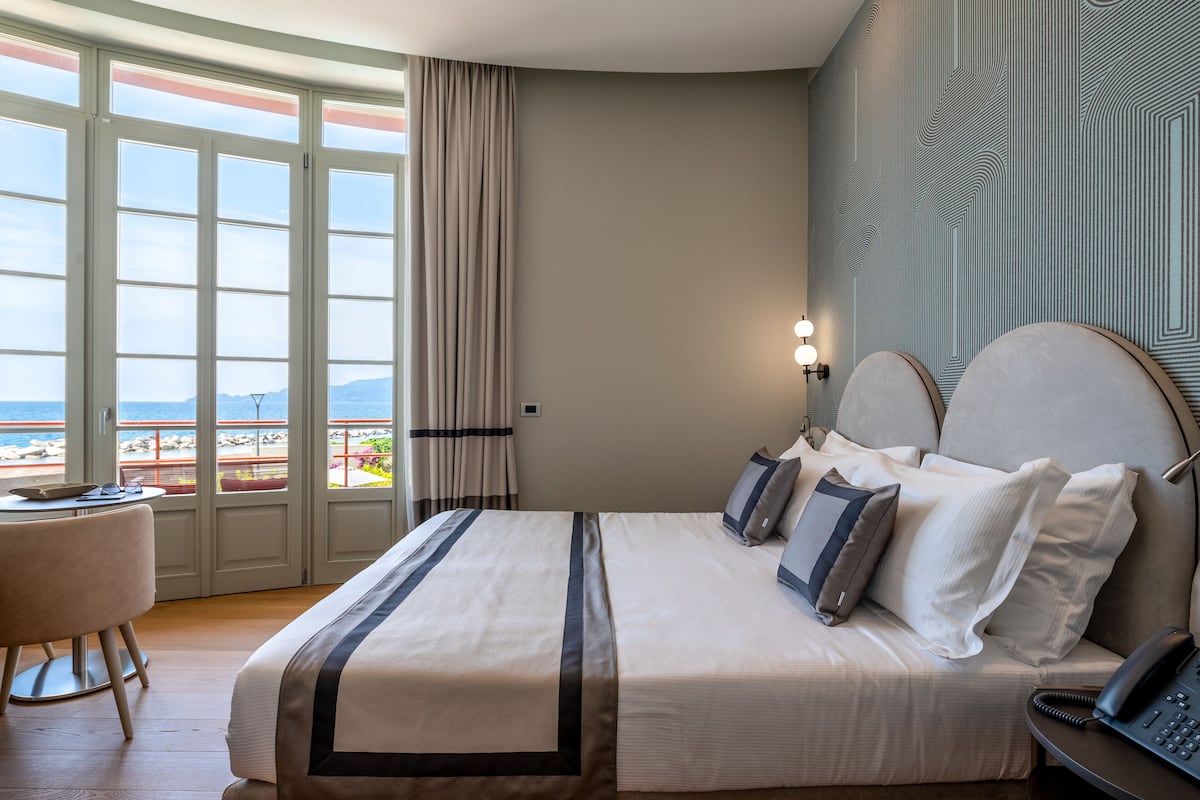A hotel room with a bed, bedside table, lamp, and a view of the beach through large windows.