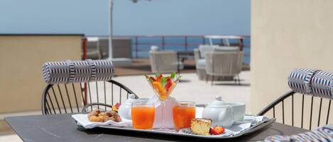 Bandeja de desayuno con jugo, ensalada de frutas y pasteles en una terraza con sillas de mimbre y vista al mar.