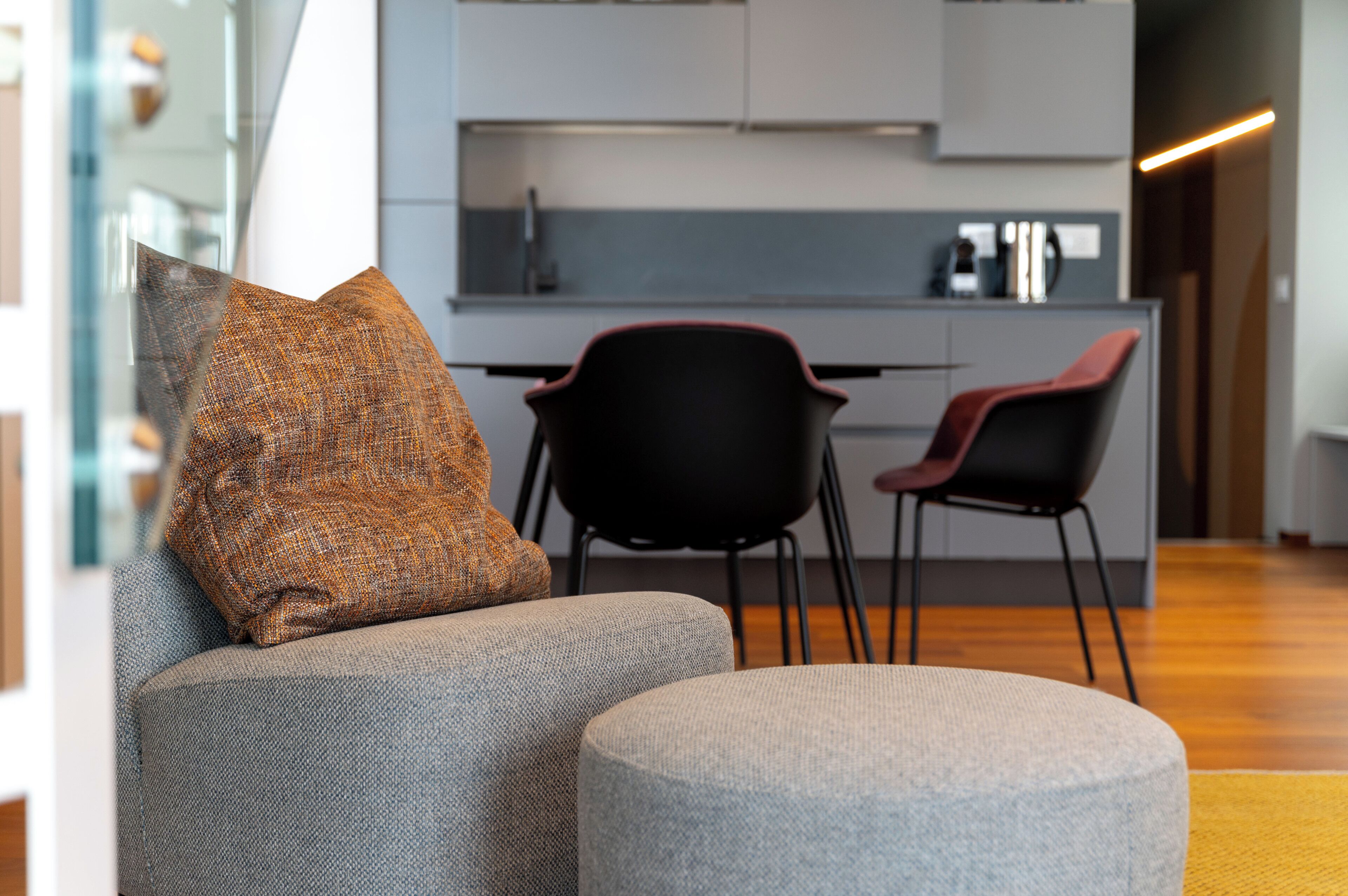 Una sala moderna con un sofá gris, un reposapiés a juego y una silla roja. Se ve una cocina con encimera gris y sillas negras al fondo.