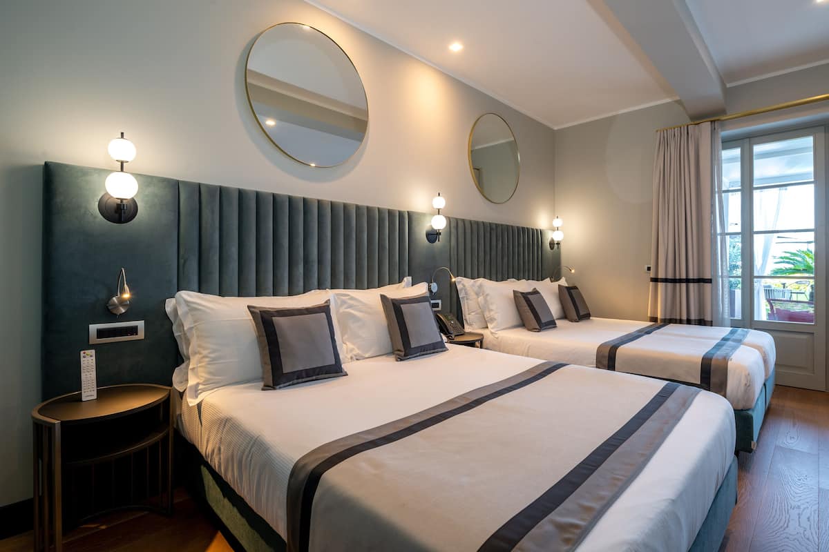 A hotel room with two beds, a large mirror, wall sconces, and a window with curtains.
