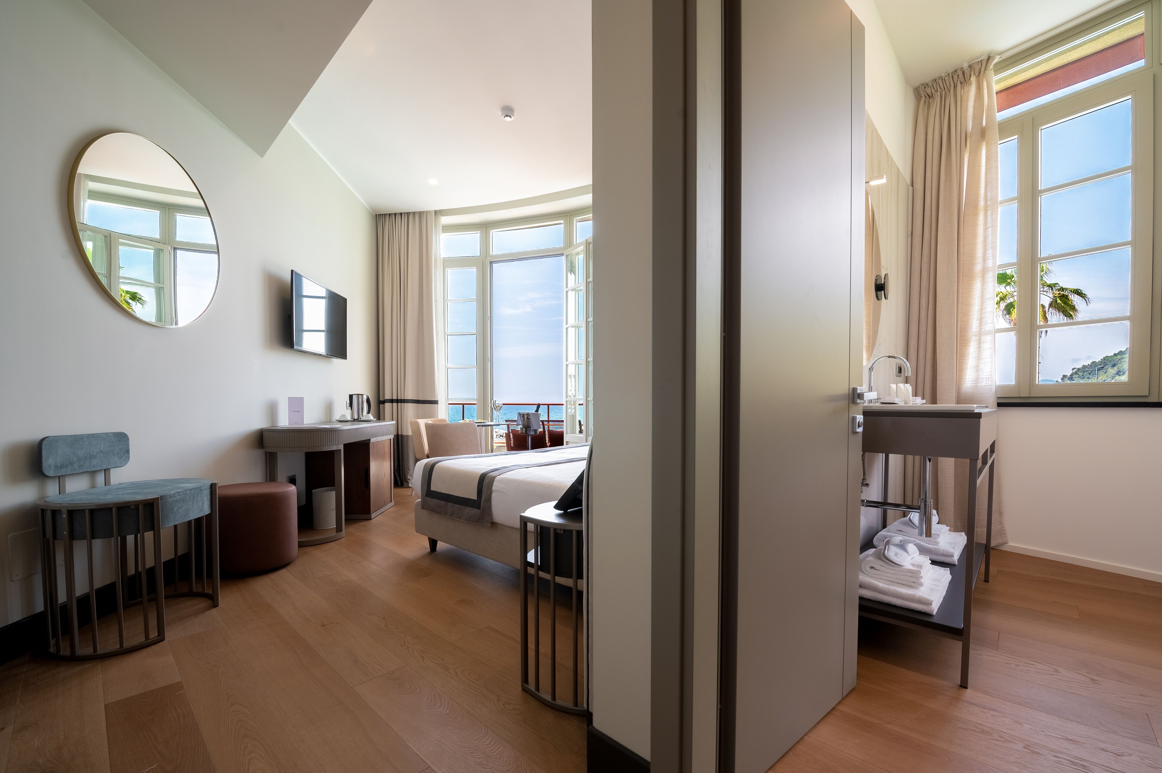 A modern hotel room with a bed, a desk, a chair, a round mirror, and a view of the sea.