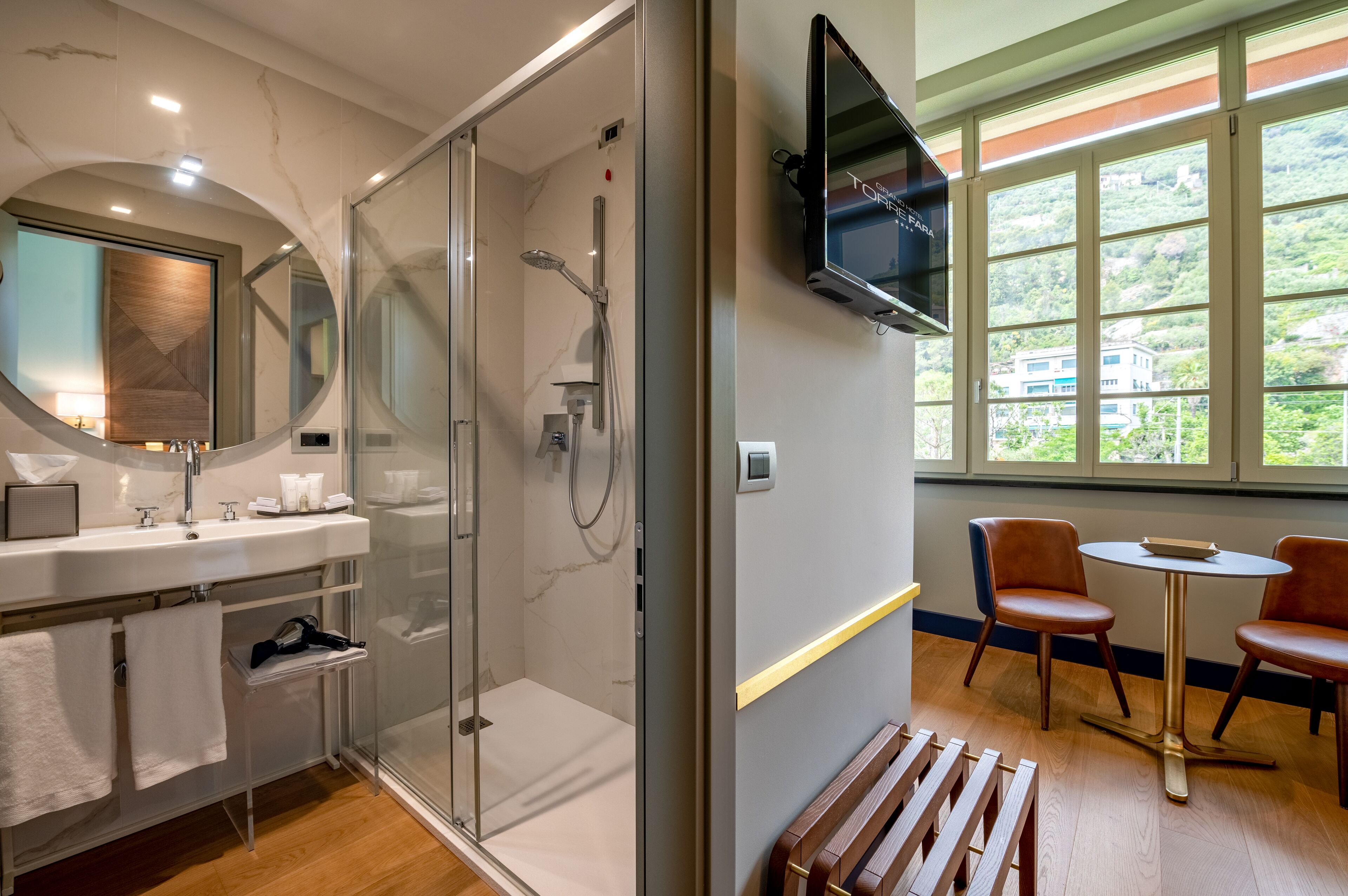 Een moderne badkamer met een glazen douchecabine, een wastafel met spiegel en een televisie boven een raam met uitzicht op groen.