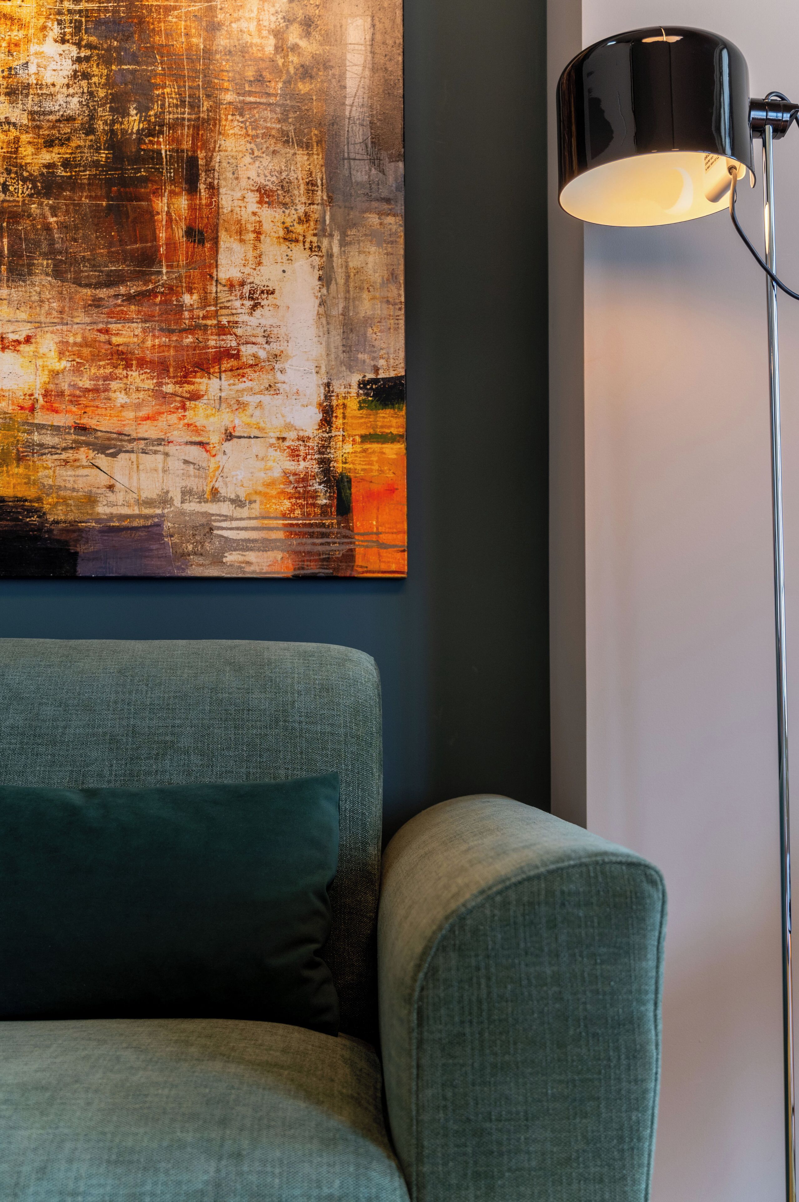Una habitación con un sillón verde, una lámpara de pie negra y un cuadro abstracto grande en la pared.