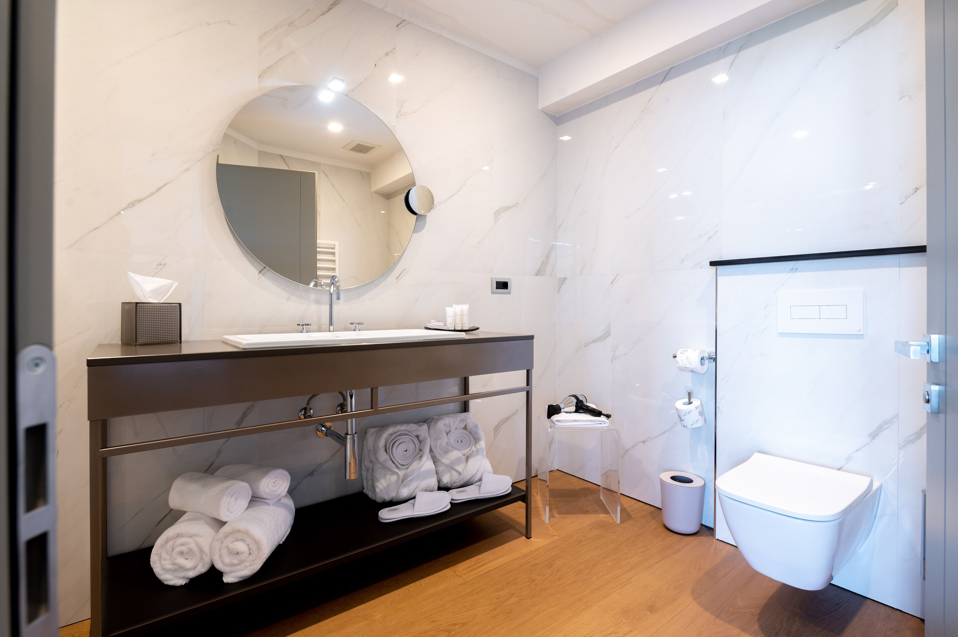 Un baño moderno con pared de mármol, espejo redondo, lavamanos, inodoro y un pequeño cubo de basura.