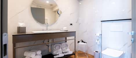 Un baño moderno con pared de mármol, espejo redondo, lavamanos, inodoro y un pequeño cubo de basura.