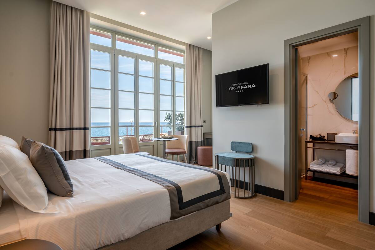 A hotel room with a large bed, a TV, a bathroom with a mirror, and a view of the sea.