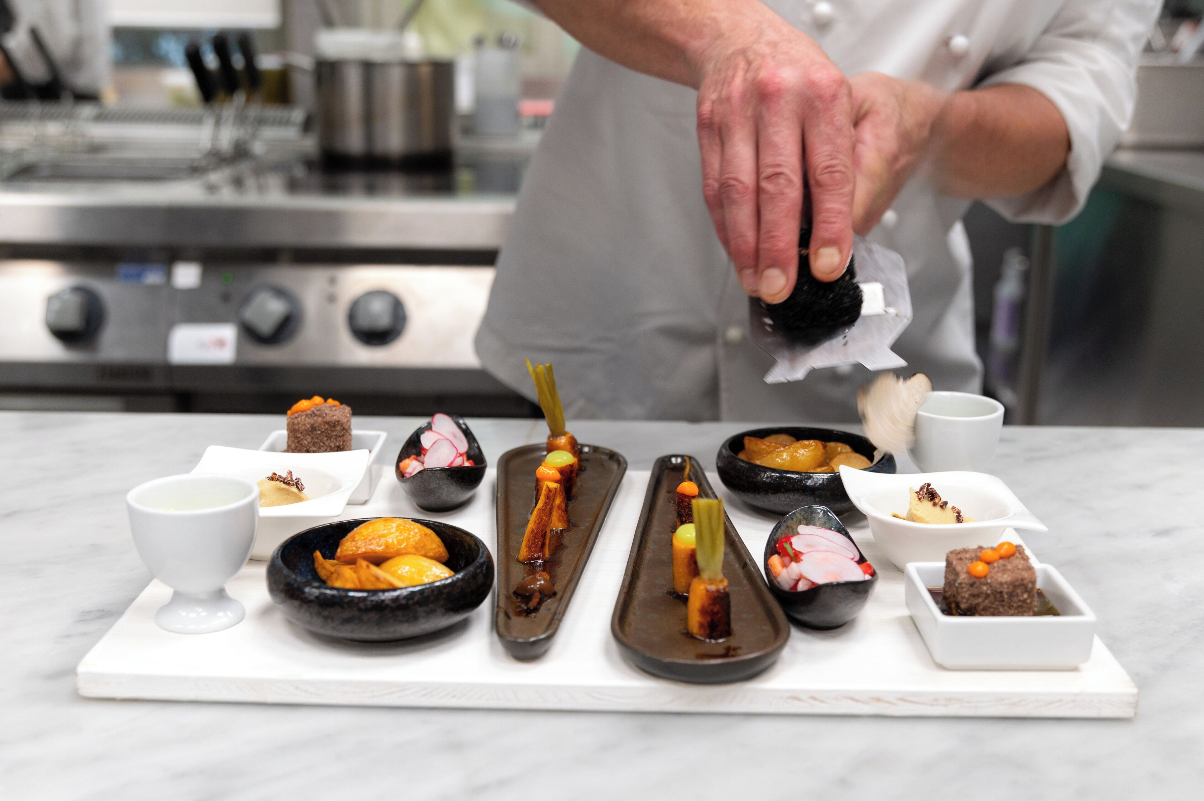 A chef preparing a dish with a focus on a variety of small, intricately arranged food items on a white serving board.