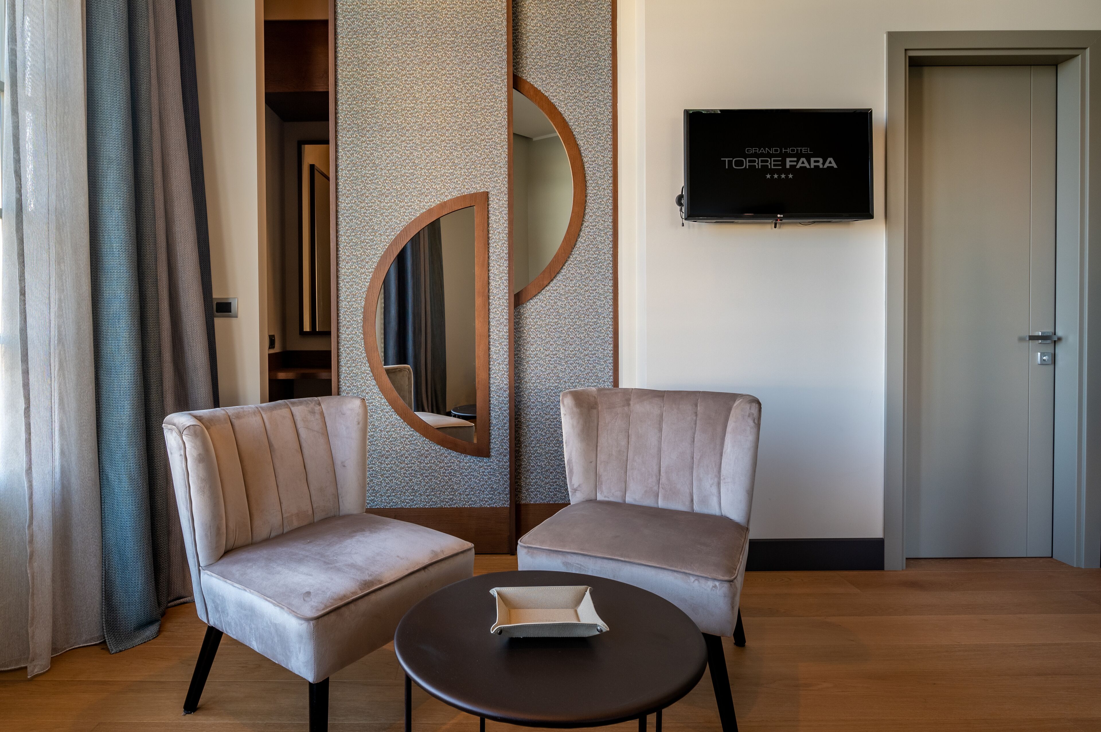Une chambre d’hôtel avec deux fauteuils, une petite table, un téléviseur fixé au mur et une porte.