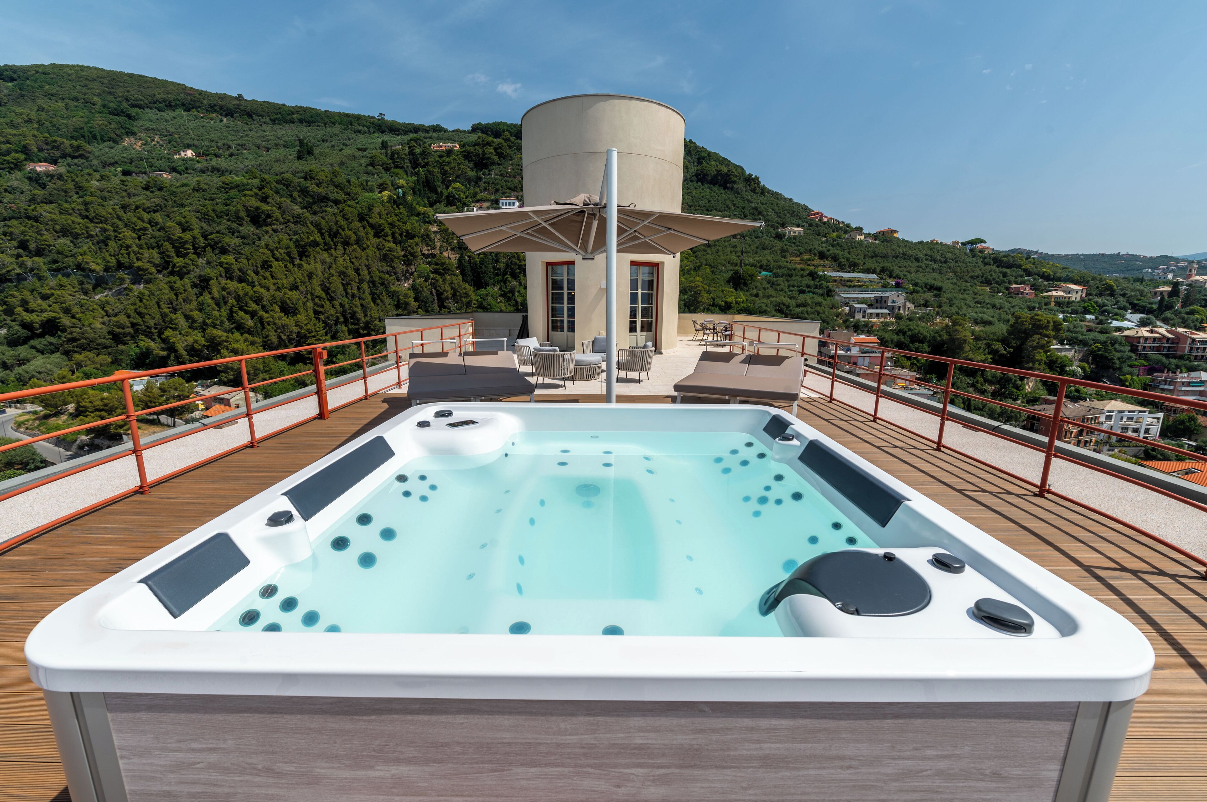 A rooftop pool with a hot tub, surrounded by a wooden deck and a red railing, overlooking a hillside with buildings.