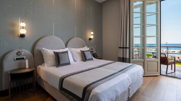 Quarto de hotel com uma cama grande, uma mesa de cabeceira, uma luminária na parede e vista para o mar através de uma porta de vidro.