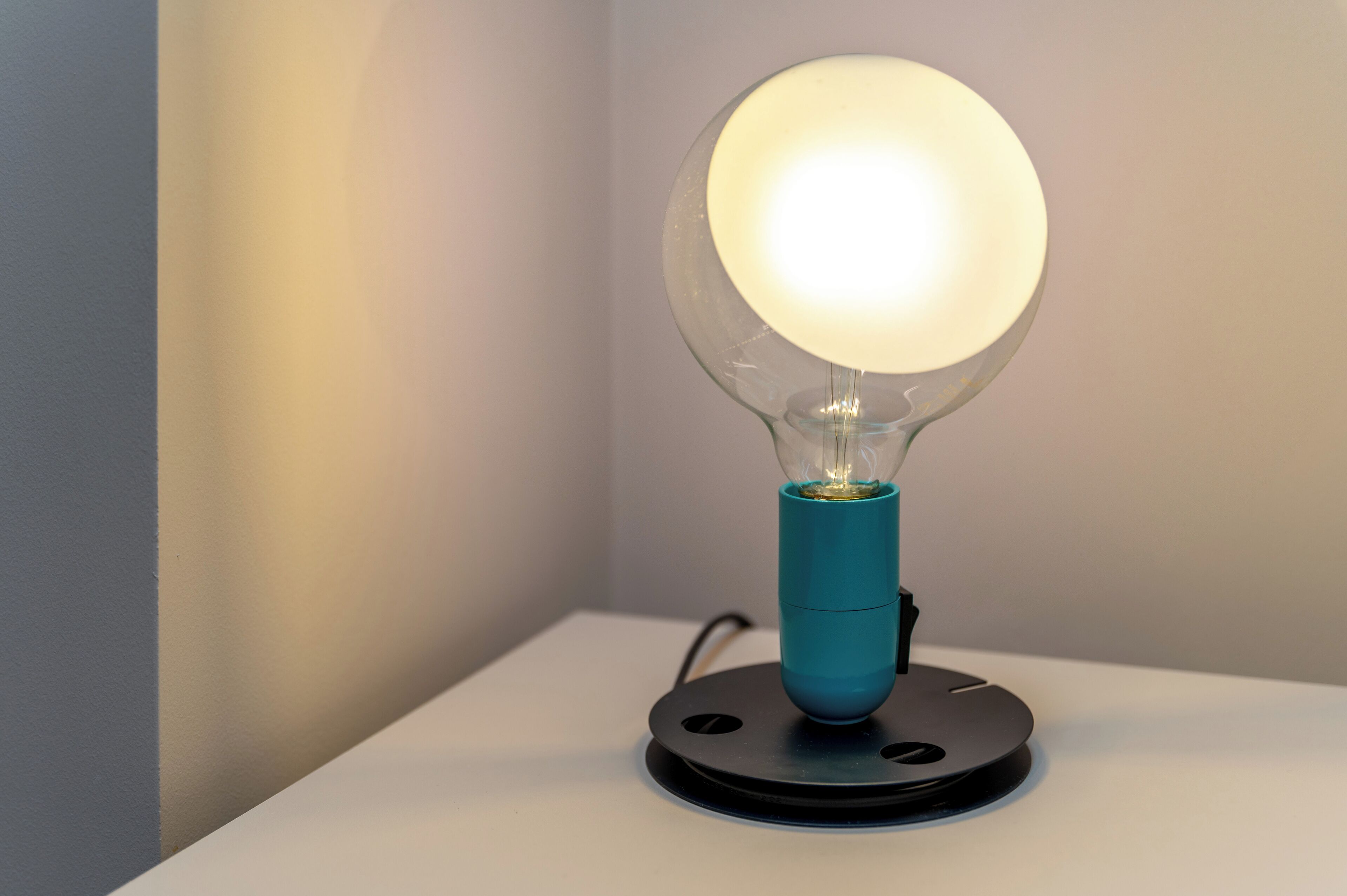 Uma luminária moderna com lâmpada de vidro transparente e base azul-turquesa sobre uma superfície branca.