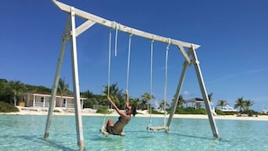 A person swinging on a swing over clear turquoise water, with a beach and resort buildings in the background. - Bahamas Luxury Sailing Catamaran, Nassau to Exumas (Staniel Cay)