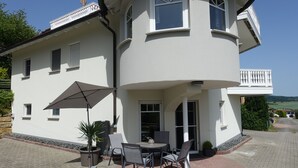 A white building with a balcony, a patio with a table and chairs, and a potted plant. - Vacation apartment RhönPanorama EG (Kaltennordheim)
