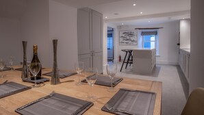 A modern dining area with a wooden table set for a meal, a kitchen in the background, and a view of the sea through a window. - Caleb's Cottage - 4 Bedroom Cottage - Solva (Haverfordwest)